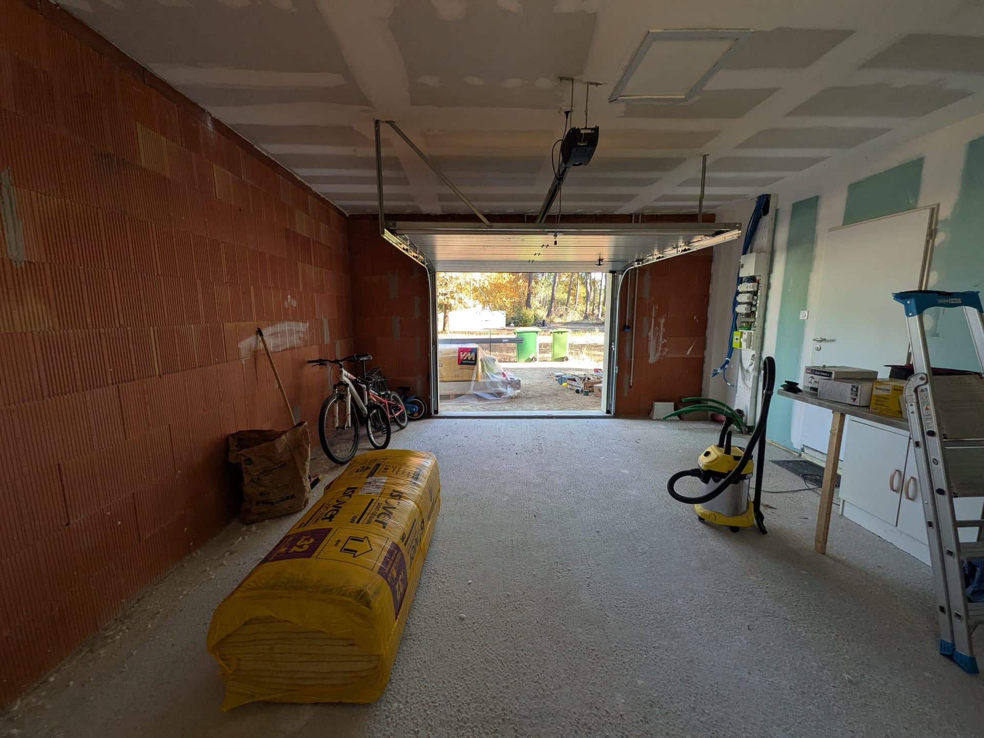 Garage avant travaux vu vers la porte sectionnelle, murs en brique nue et rouleaux de laine de verre Isover GR32 au sol